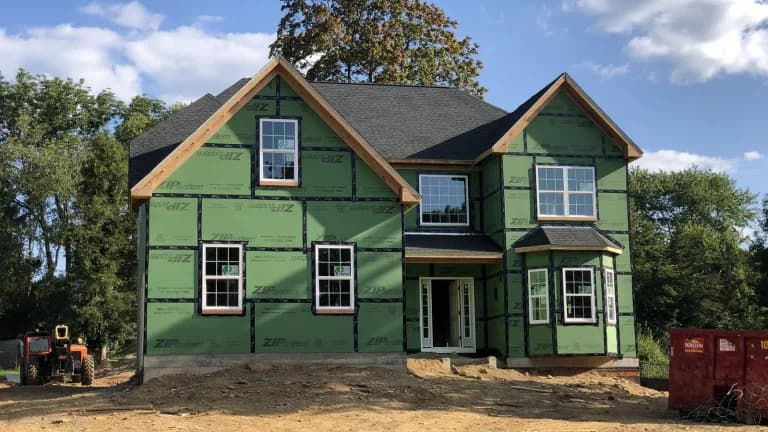 A two-story new home wrapped in green sheathing with windows installed, on a dirt lot with a small tractor and a dumpster
