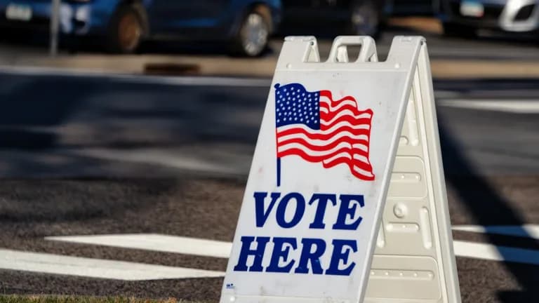 A white sidewalk sign with an American flag and the words Vote Here on a grassy curb, with parked cars blurred behind it