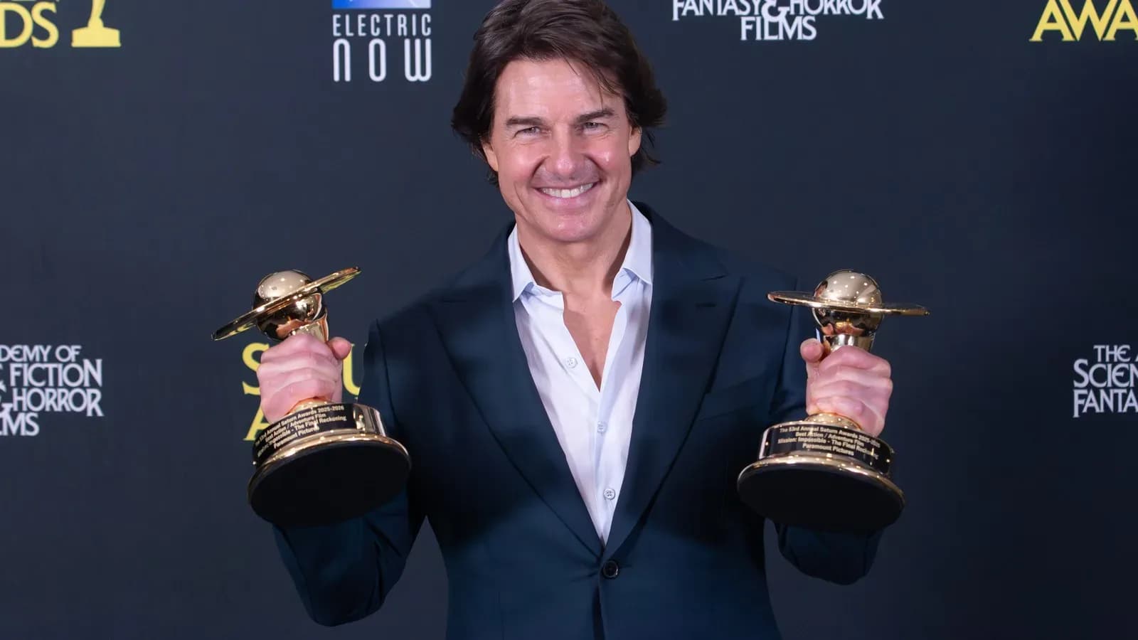 Tom Cruise in a dark blue suit smiling and holding two gold Saturn Award trophies in front of a black backdrop
