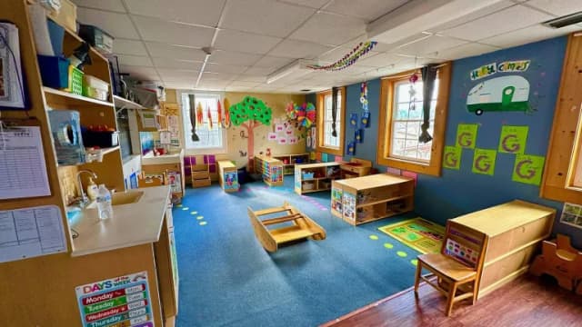 An empty child care classroom with small tables, shelves and a blue rug