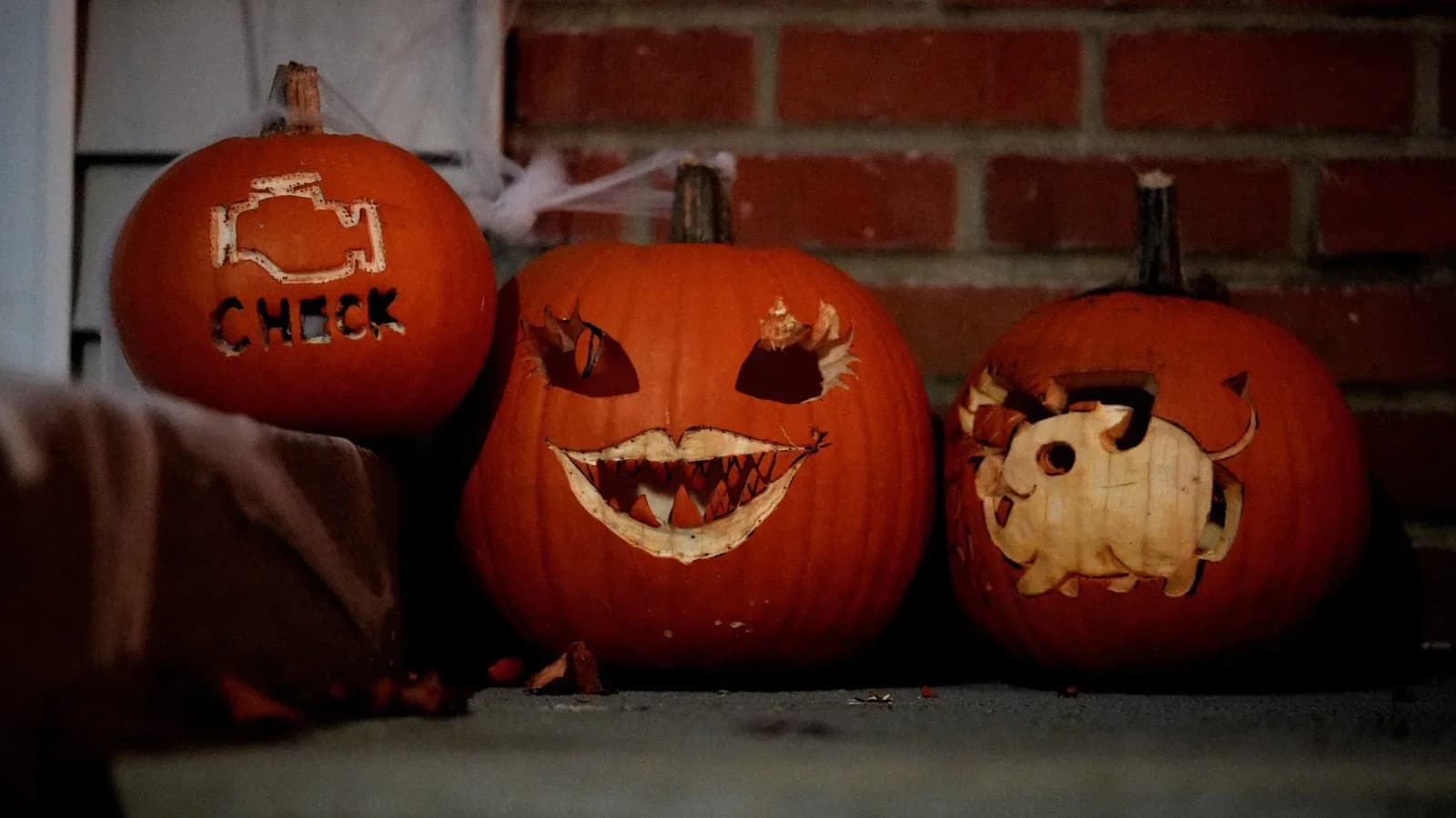 Three carved jack-o'-lanterns sitting on a porch beside a brick wall at dusk