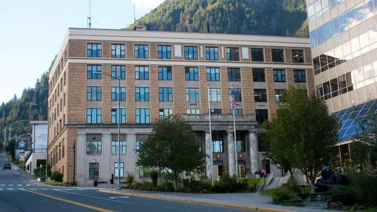 The Alaska State Capitol, a brick and stone office building with a marble-columned entrance, on a downtown street below a forested mountain in Juneau