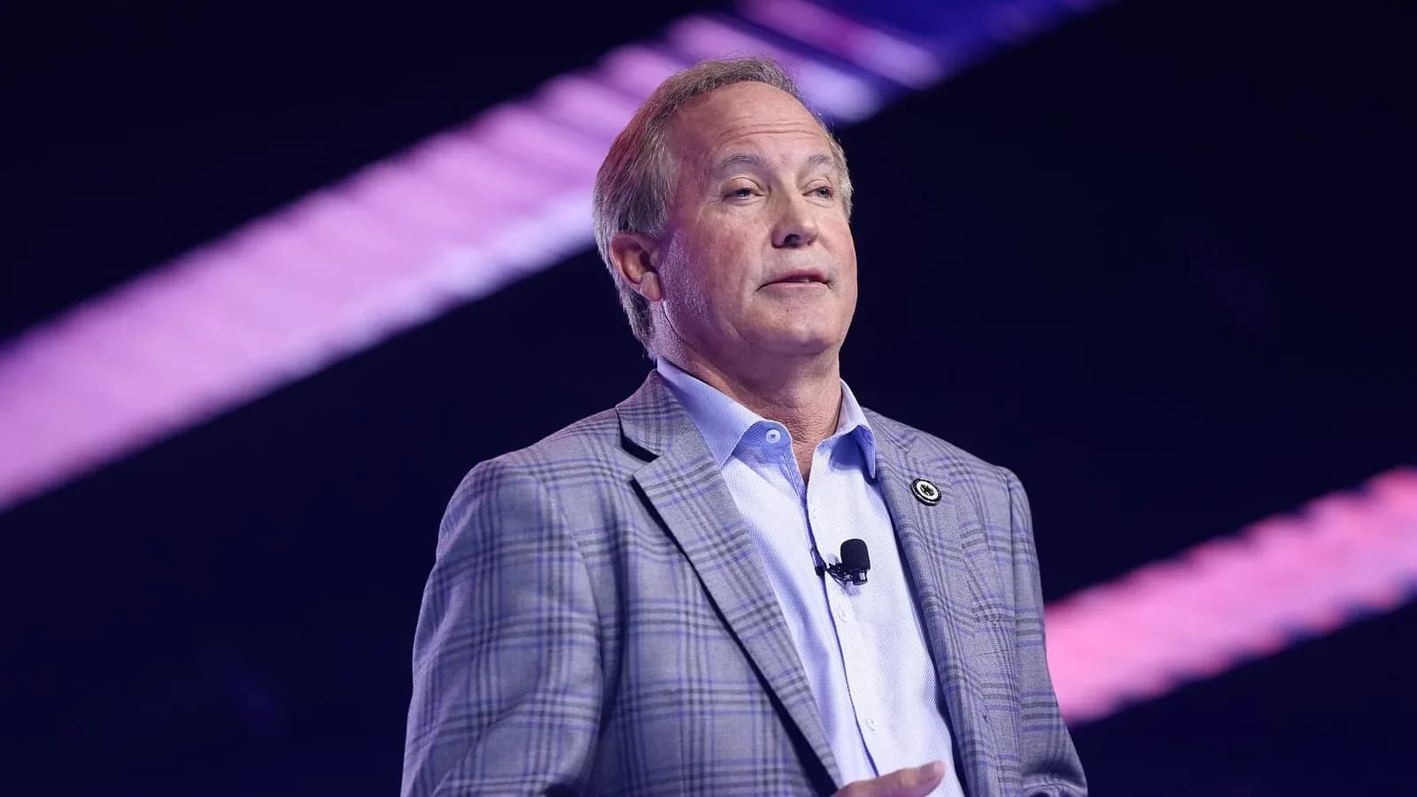 Texas Attorney General Ken Paxton, in a plaid blazer, speaks on a stage with purple lighting