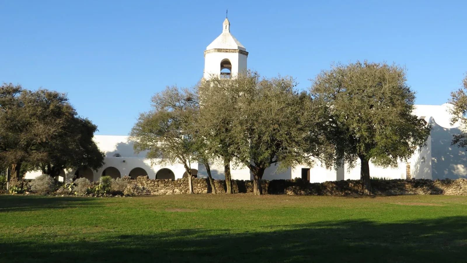 The whitewashed Mission Espíritu Santo church and bell tower behind live oaks in Goliad, Texas