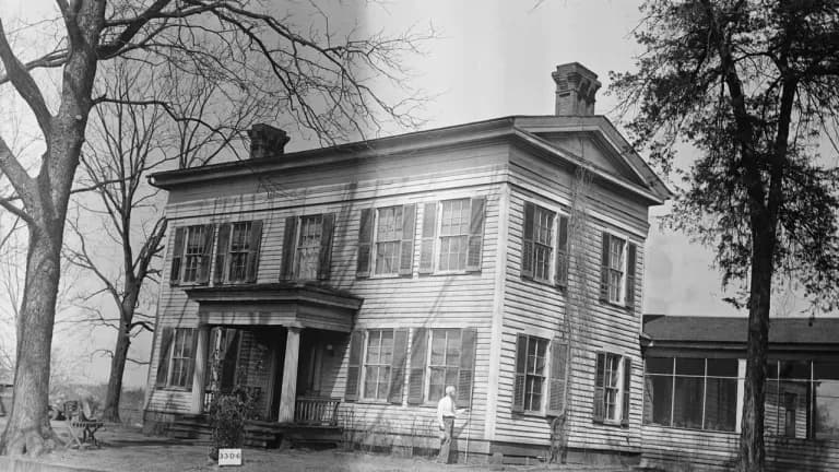 Black-and-white 1934 photograph of the Matthew Cartwright House in San Augustine, Texas, a two-story white clapboard Greek Revival house with dark shutters, a small columned entrance porch, two brick chimneys and a low pediment, framed by tall bare trees, with a man standing beside the front wall