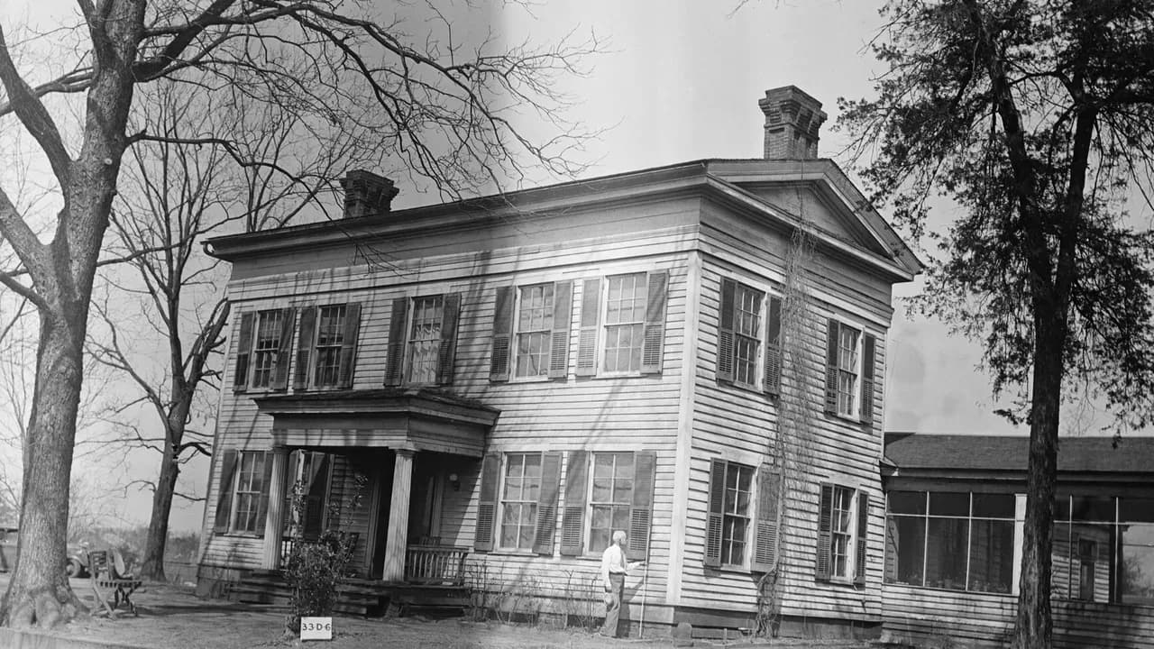 Black-and-white 1934 photograph of the Matthew Cartwright House in San Augustine, Texas, a two-story white clapboard Greek Revival house with dark shutters, a small columned entrance porch, two brick chimneys and a low pediment, framed by tall bare trees, with a man standing beside the front wall
