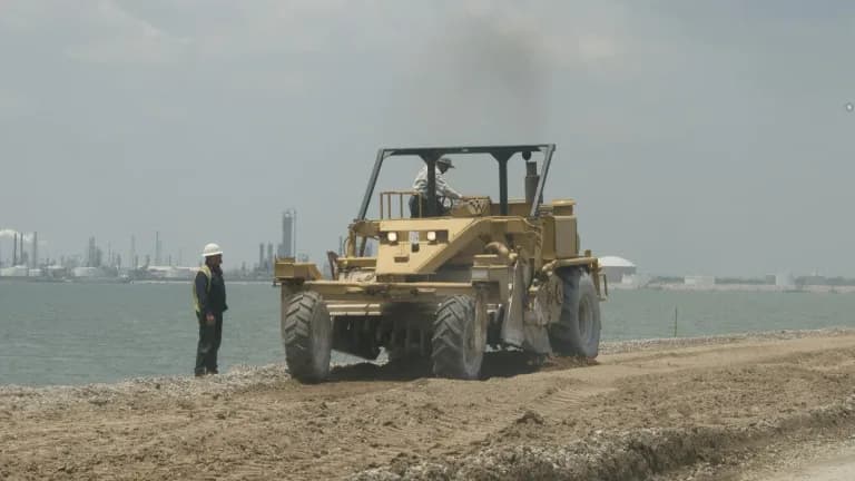 A worker in a hard hat watches a road grader rebuild a gravel roadbed beside the water