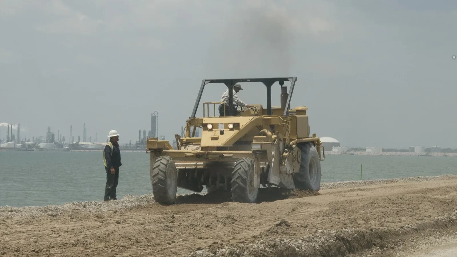 A worker in a hard hat watches a road grader rebuild a gravel roadbed beside the water