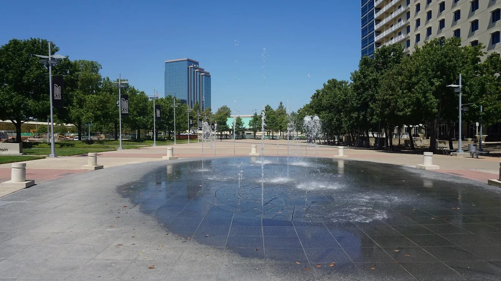A wide plaza with a round fountain pool, trees and office towers in Richardson under a blue sky