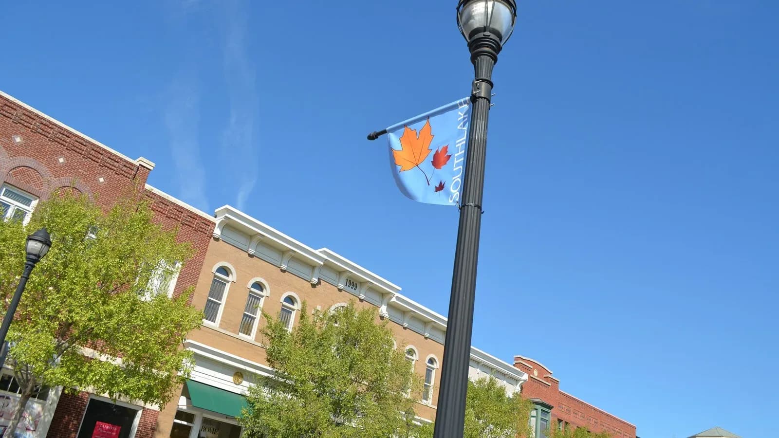 Red brick storefronts at Southlake Town Square with a lamppost and a hanging banner under a bright blue sky