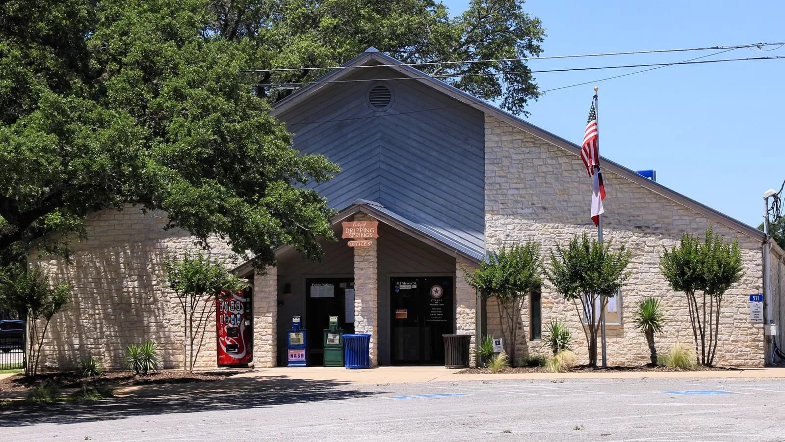 A limestone and metal-roofed Dripping Springs city building with an American flag, shade trees and a parking lot