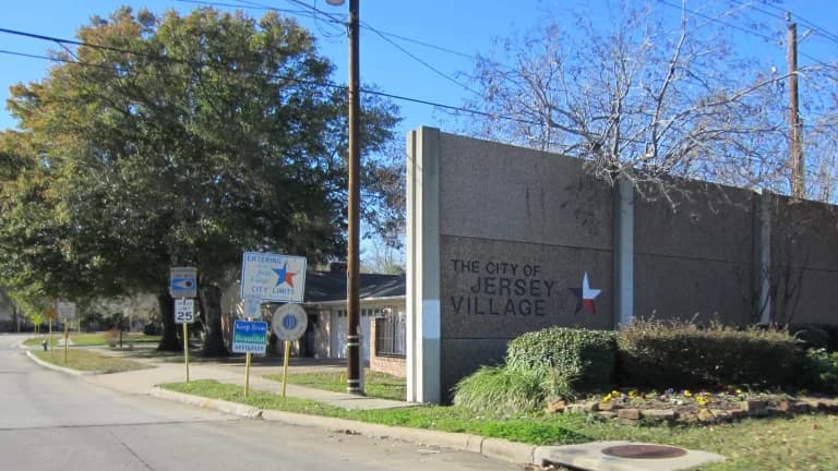 A concrete wall reading The City of Jersey Village beside a street with trees and a sidewalk