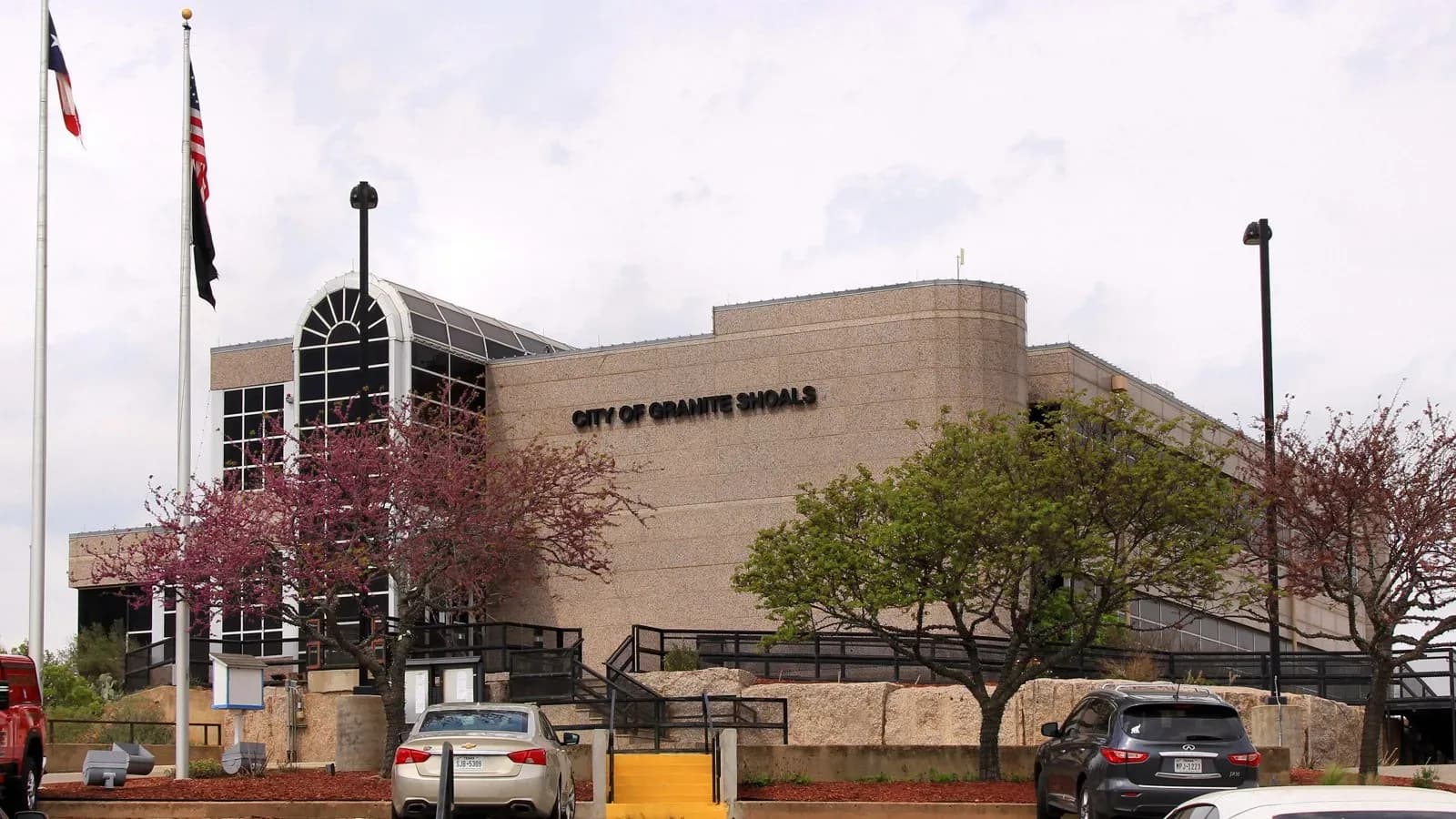 The tan Granite Shoals City Hall building with flagpoles and flowering trees out front