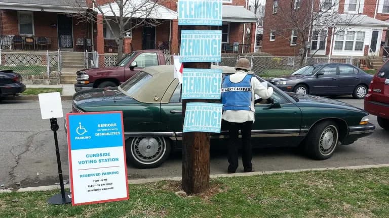 A blue curbside voting sign beside a parked car outside a polling place