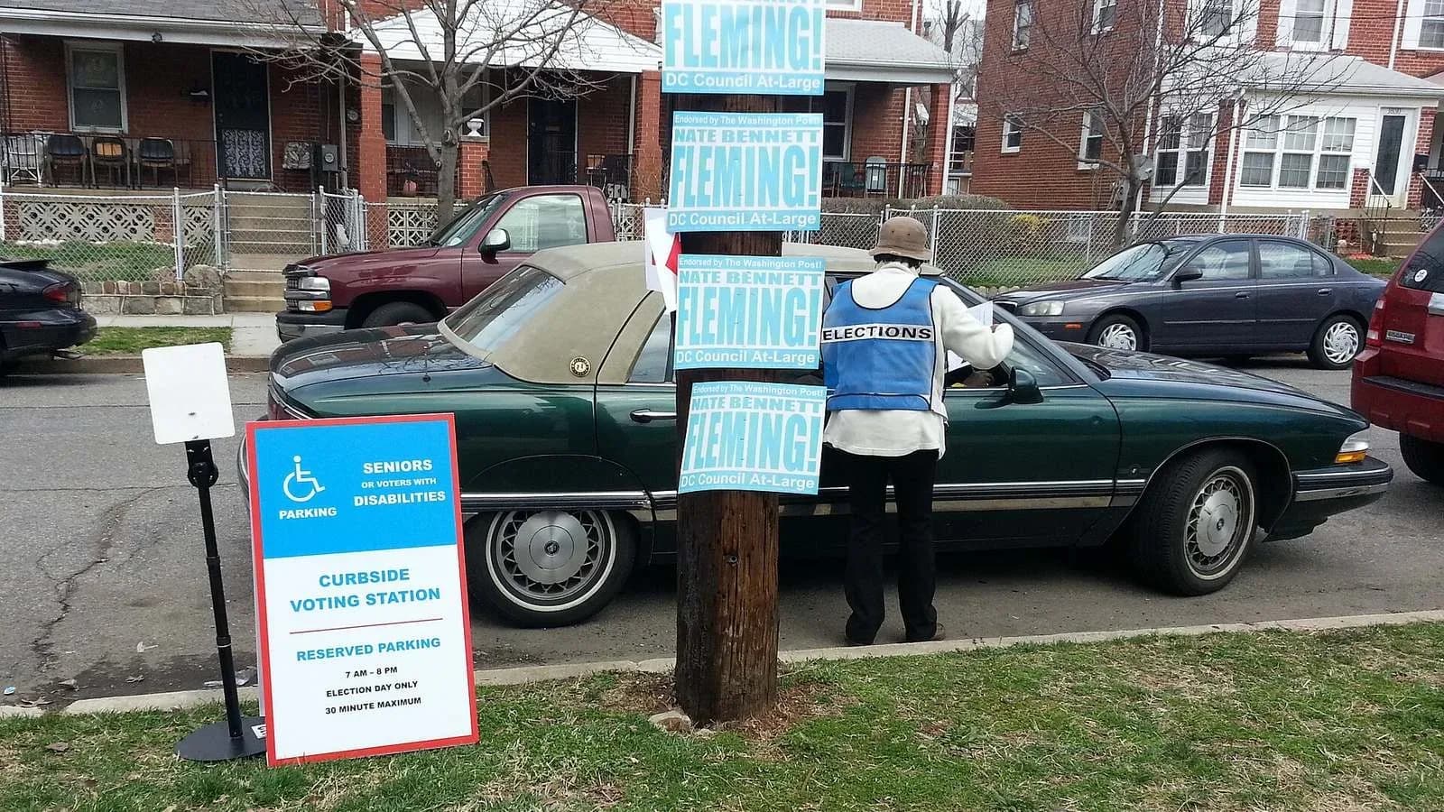 A blue curbside voting sign beside a parked car outside a polling place