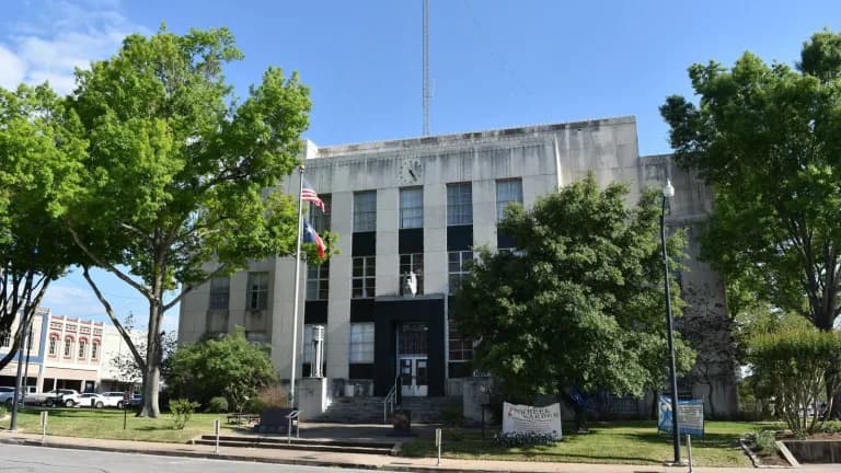 The pale stone Washington County Courthouse with American and Texas flags out front and tall trees on either side