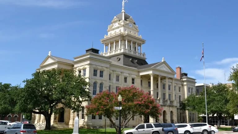 The white stone Bell County Courthouse with a clock tower and a statue on top, framed by trees under a blue sky