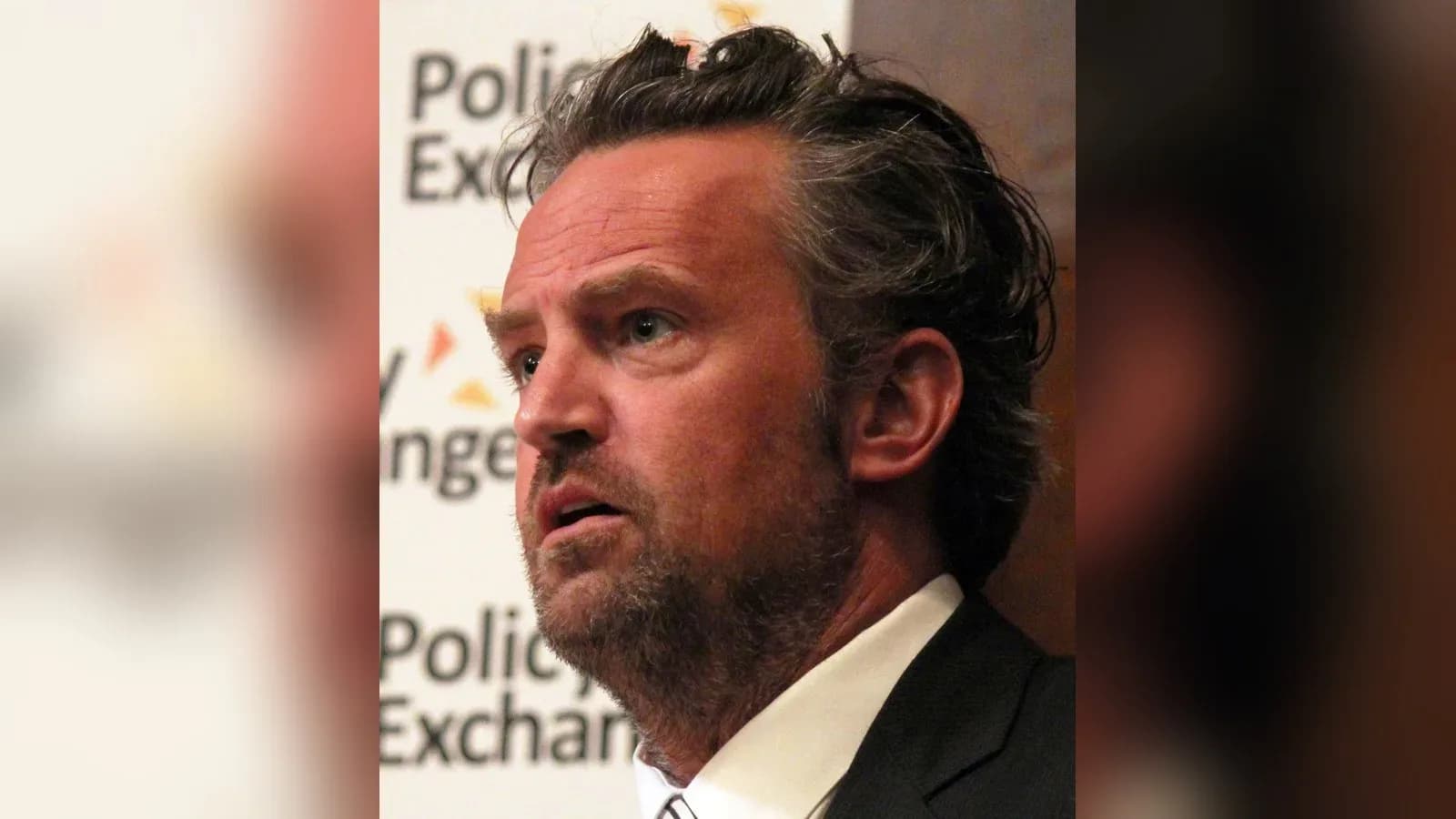 Matthew Perry speaking in front of a Policy Exchange backdrop, wearing a dark suit and striped tie