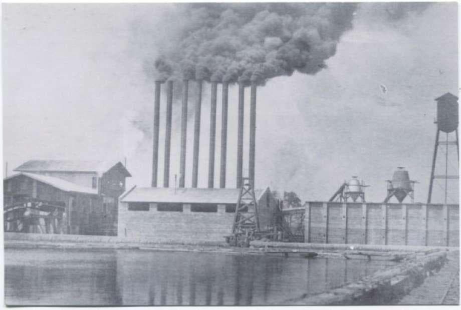 Early Conroe sawmill beside a mill pond, with a row of smokestacks rising over the Piney Woods