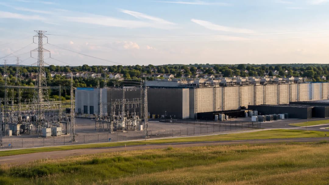 A large data center beside an electrical substation with transmission lines and nearby suburban homes
