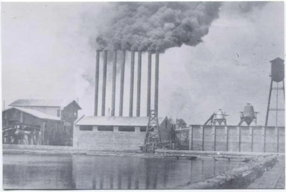 Early Conroe sawmill beside a mill pond, with a row of smokestacks rising over the Piney Woods