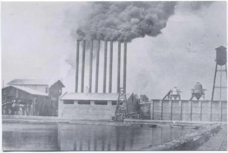 Early Conroe sawmill beside a mill pond, with a row of smokestacks rising over the Piney Woods