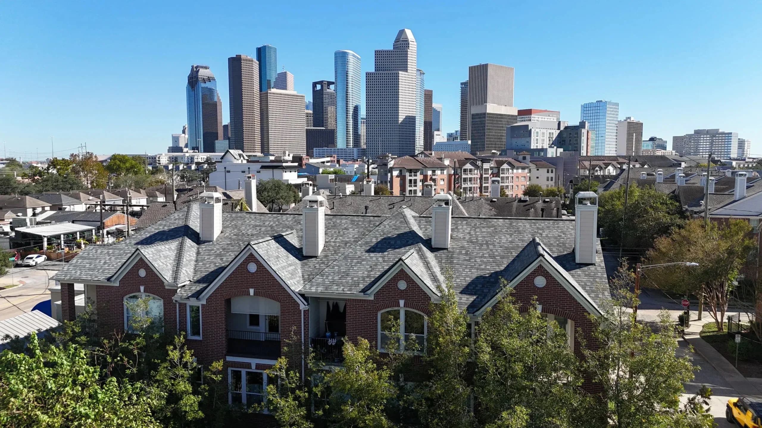 Gray architectural shingles on a brick townhome row, with the Houston skyline behind the roofs.