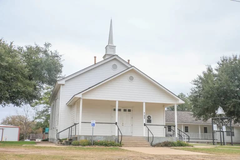 White wood-frame Plantersville Baptist Church with a steeple, a front porch, and a bell on the lawn