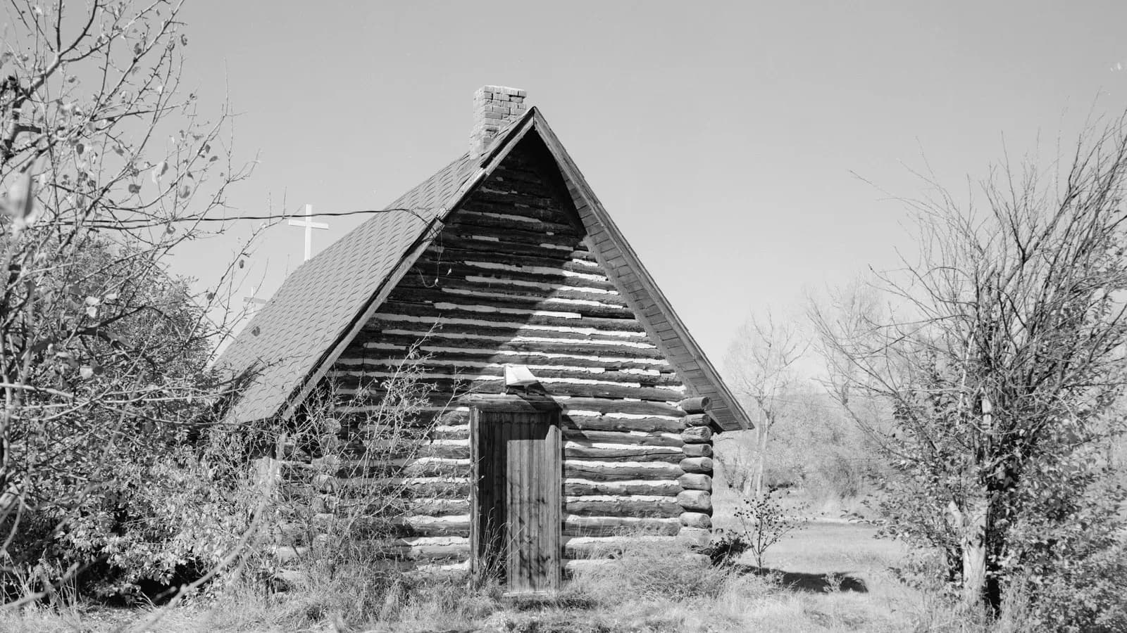 A small log chapel with a cross on its roof among trees, in black and white