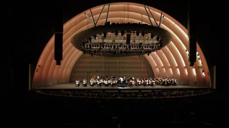 The lit arches of the Hollywood Bowl shell at night with an orchestra performing on stage and a darkened audience in front