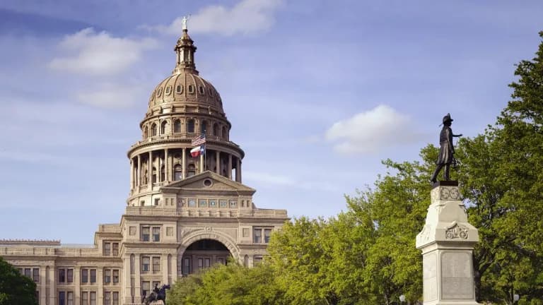 The Texas State Capitol in Austin under a blue sky, with trees and a monument on the grounds