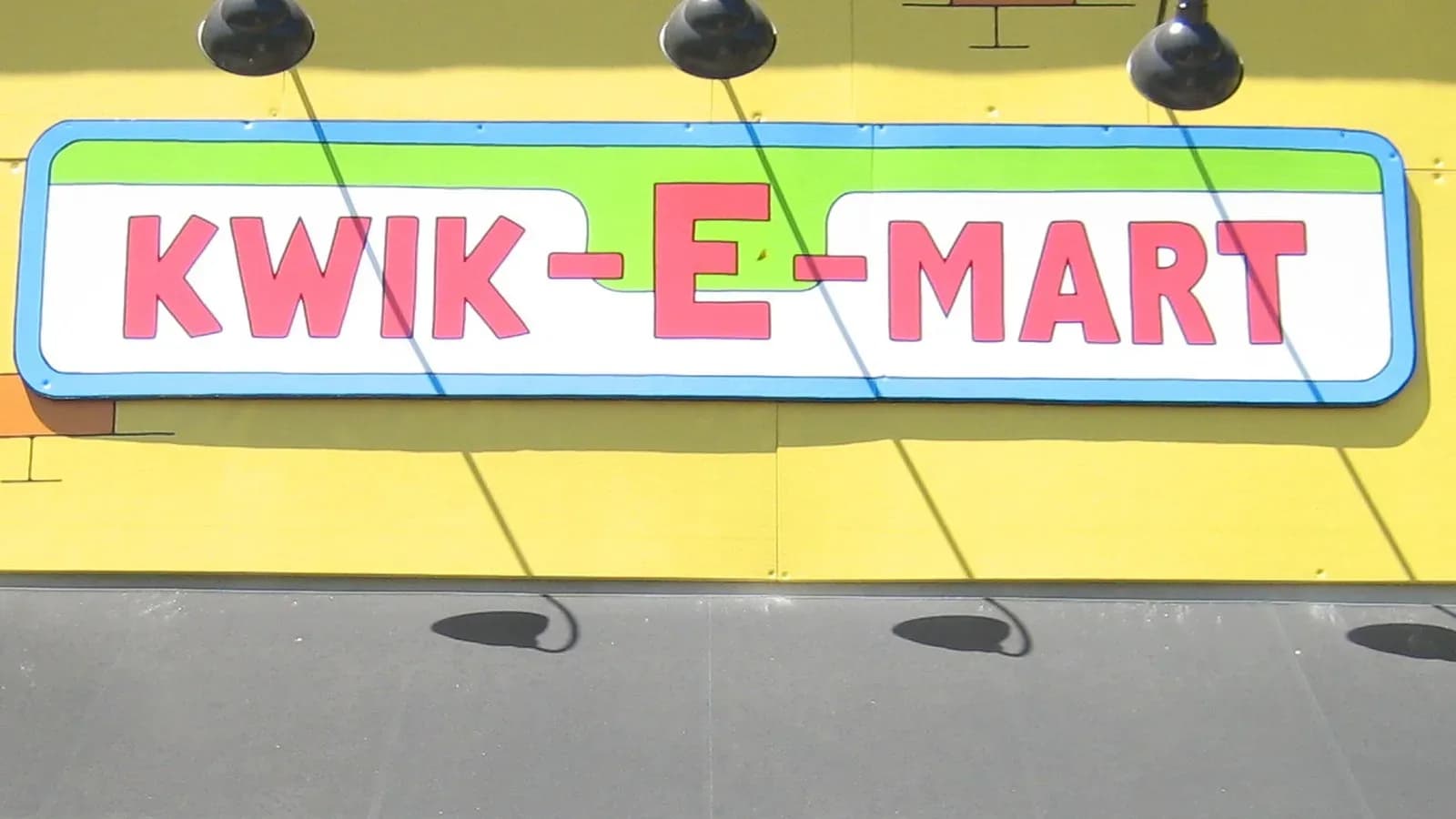 A red, green and white Kwik-E-Mart sign mounted on a bright yellow storefront wall above a gray awning