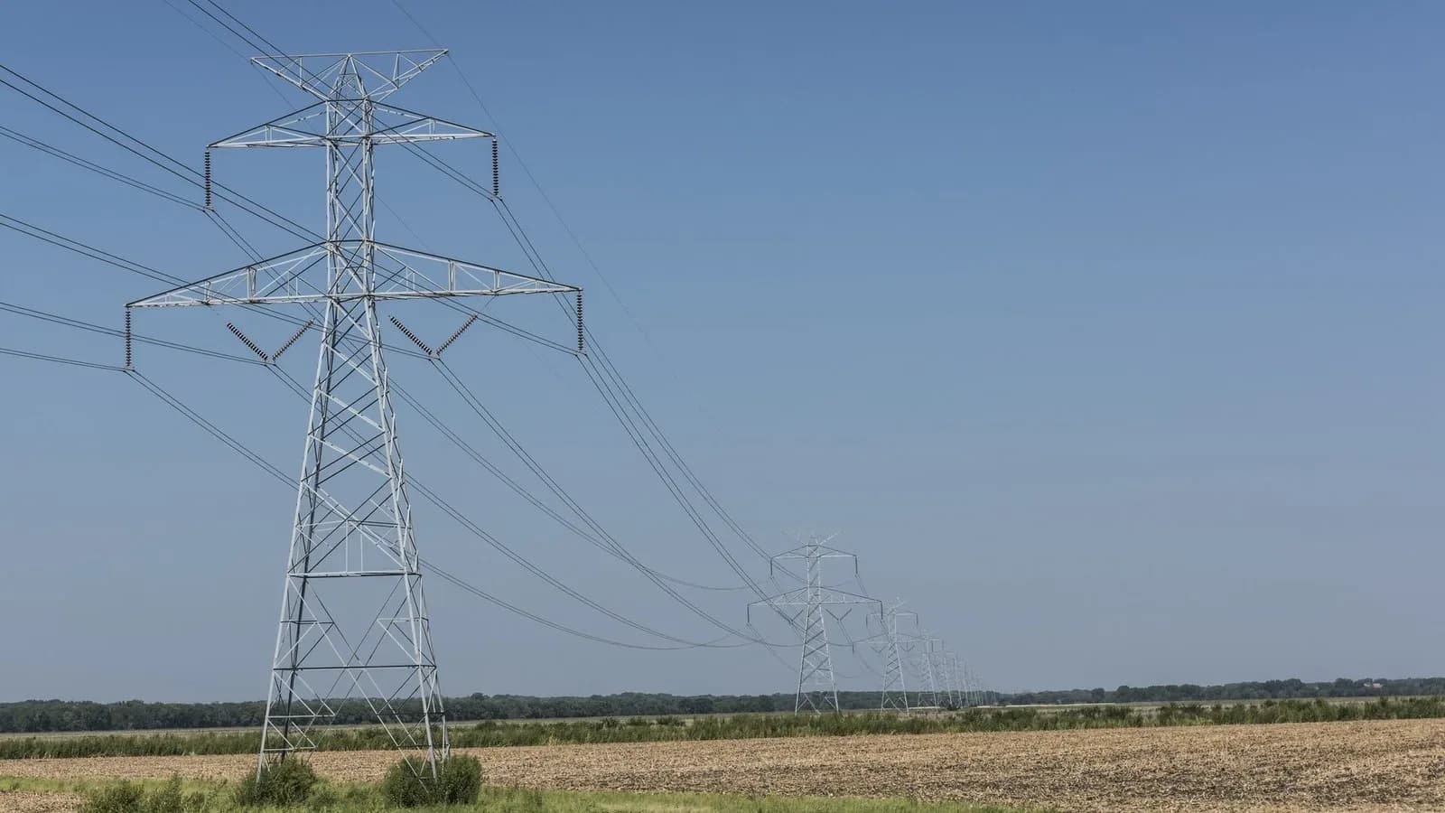 A row of steel electric transmission towers carrying power lines across flat farmland under a clear blue sky