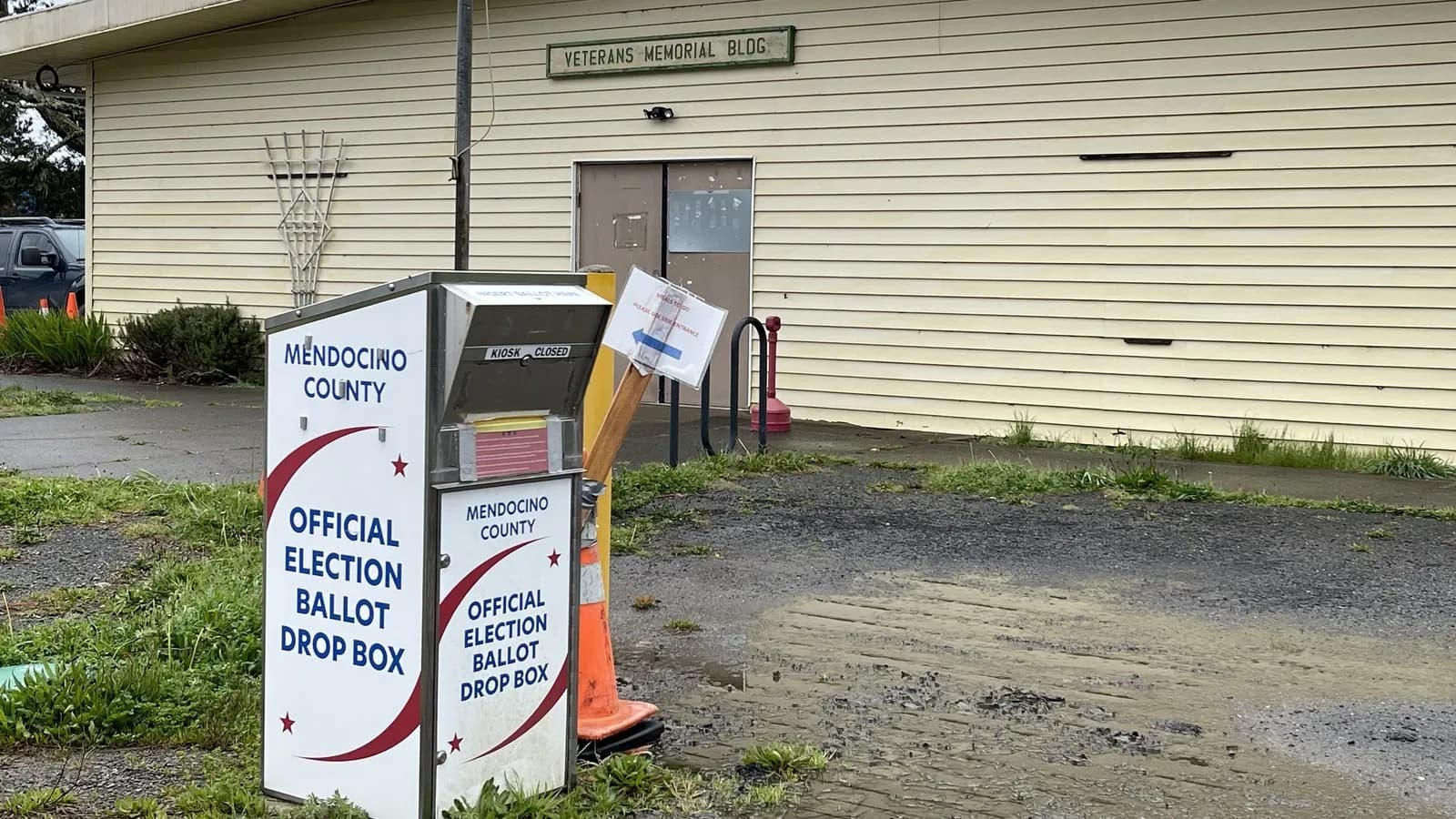 A white Mendocino County official election ballot drop box standing on a gravel lot in front of a veterans memorial building in Point Arena, California