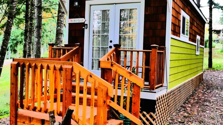 A small wooden tiny home with an orange deck and green siding among trees