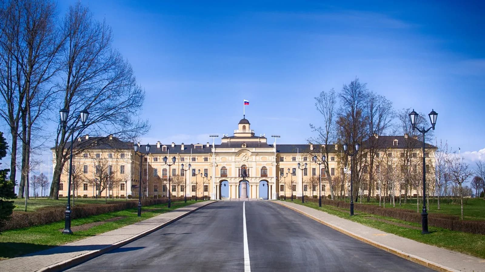 A long yellow palace with a central dome and Russian flag at the end of a straight paved road lined with lamps and bare trees