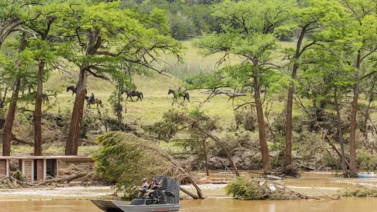 An airboat and riders on horseback along a flood-damaged riverbank in Kerrville