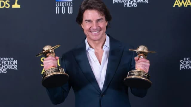 Tom Cruise in a dark blue suit smiling and holding two gold Saturn Award trophies in front of a black backdrop