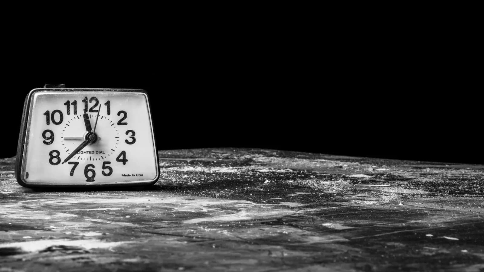 A white-faced analog alarm clock sitting on a worn tabletop against a black background, in black and white