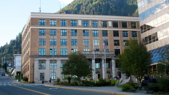 The Alaska State Capitol, a brick and stone office building with a marble-columned entrance, on a downtown street below a forested mountain in Juneau