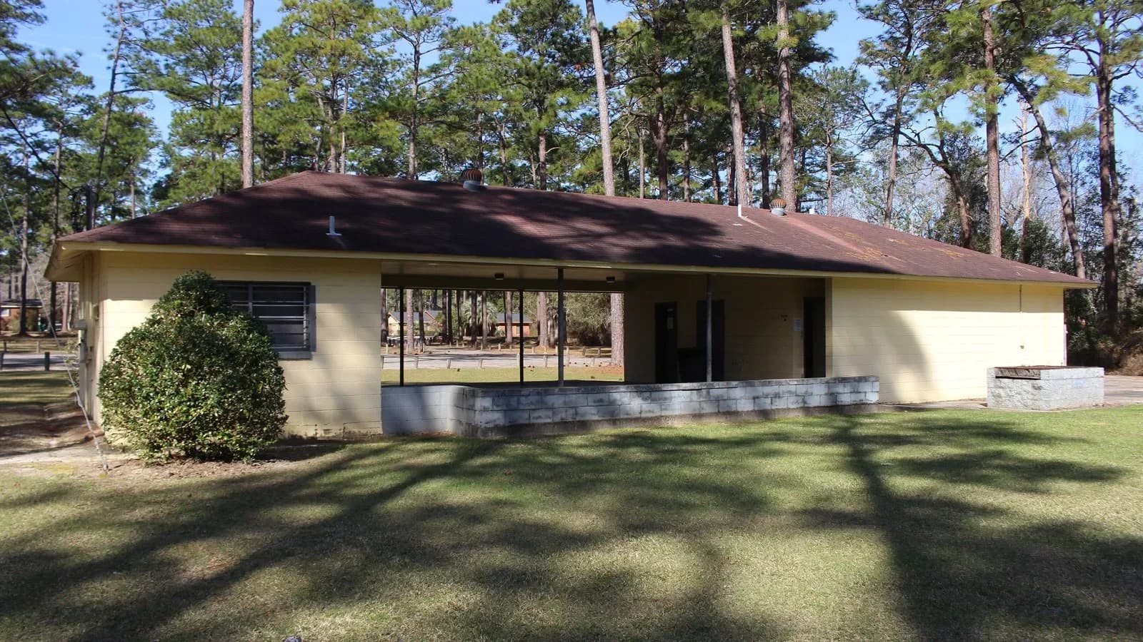 A tan brick restroom building in a park shaded by pine trees