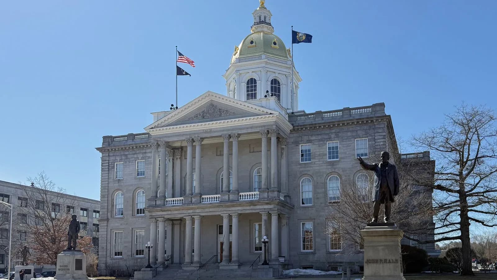 The granite New Hampshire State House in Concord with a gold dome, columned portico and bronze statues on the lawn under a blue sky