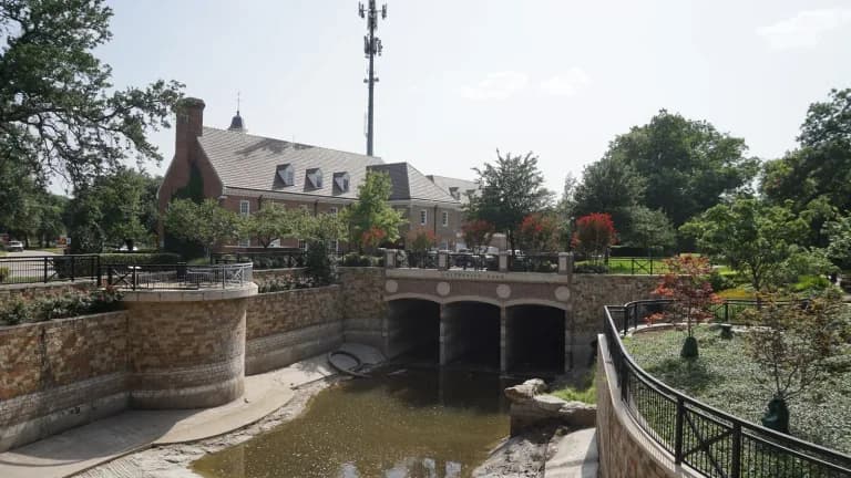 University Park City Hall, a red-brick building with dormers, behind a stone-walled creek channel and bridge