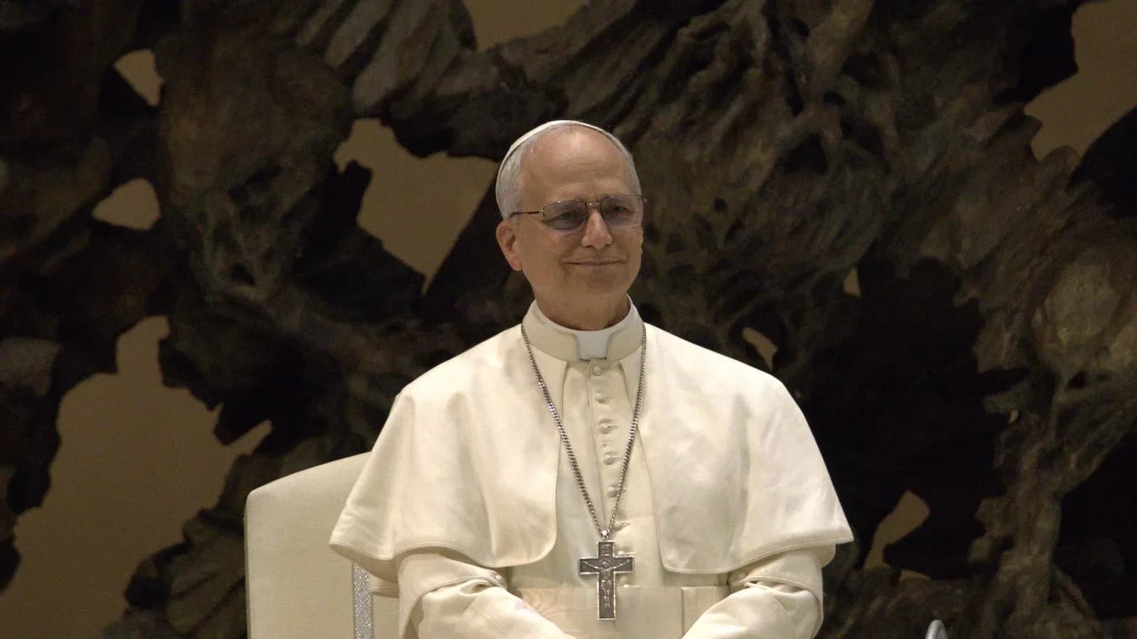 Pope Leo XIV in white cassock and pectoral cross, smiling, seated in the Paul VI Audience Hall