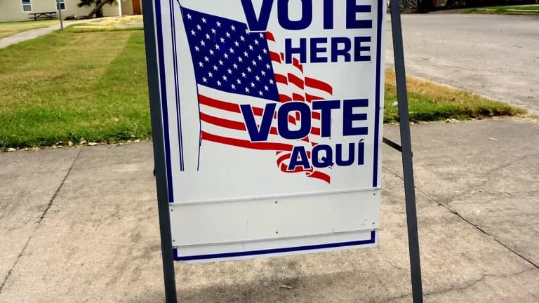 A Vote Here and Vote Aqui sign with an American flag design stands on a sidewalk outside a polling place