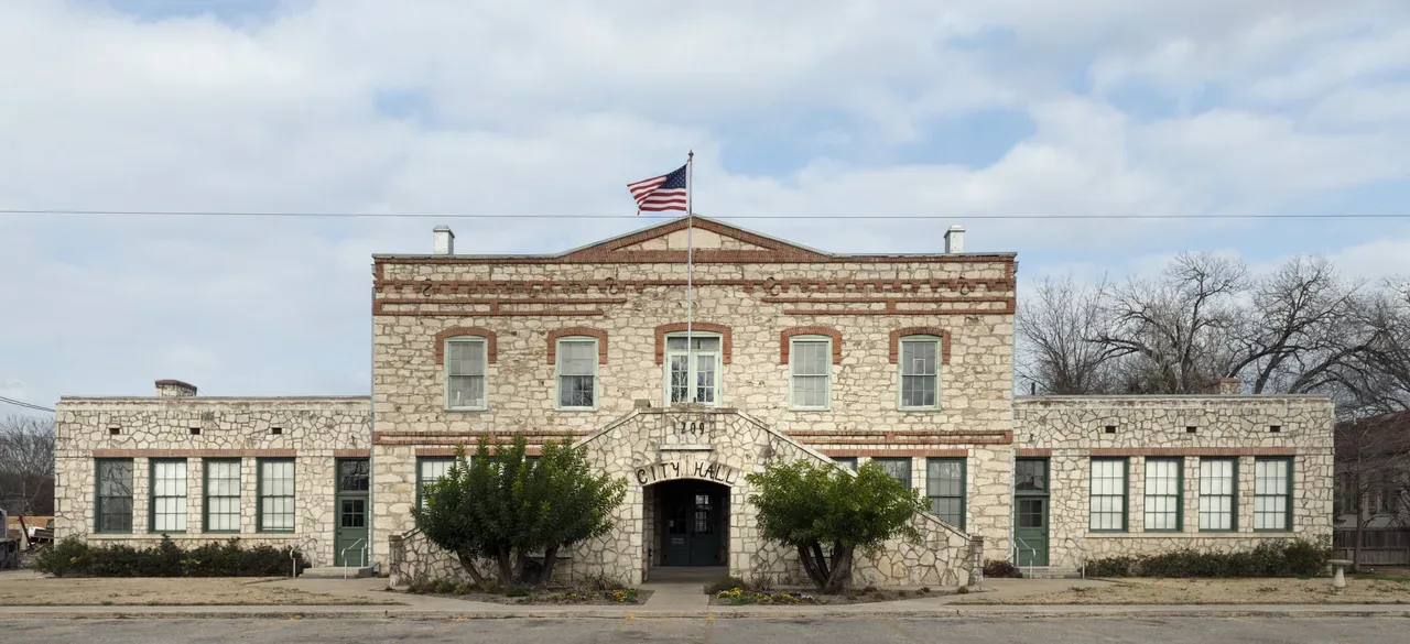 Two-story limestone building with a gabled center, flanking one-story wings and a stone entry stair marked City Hall, with an American flag, in Castroville, Texas