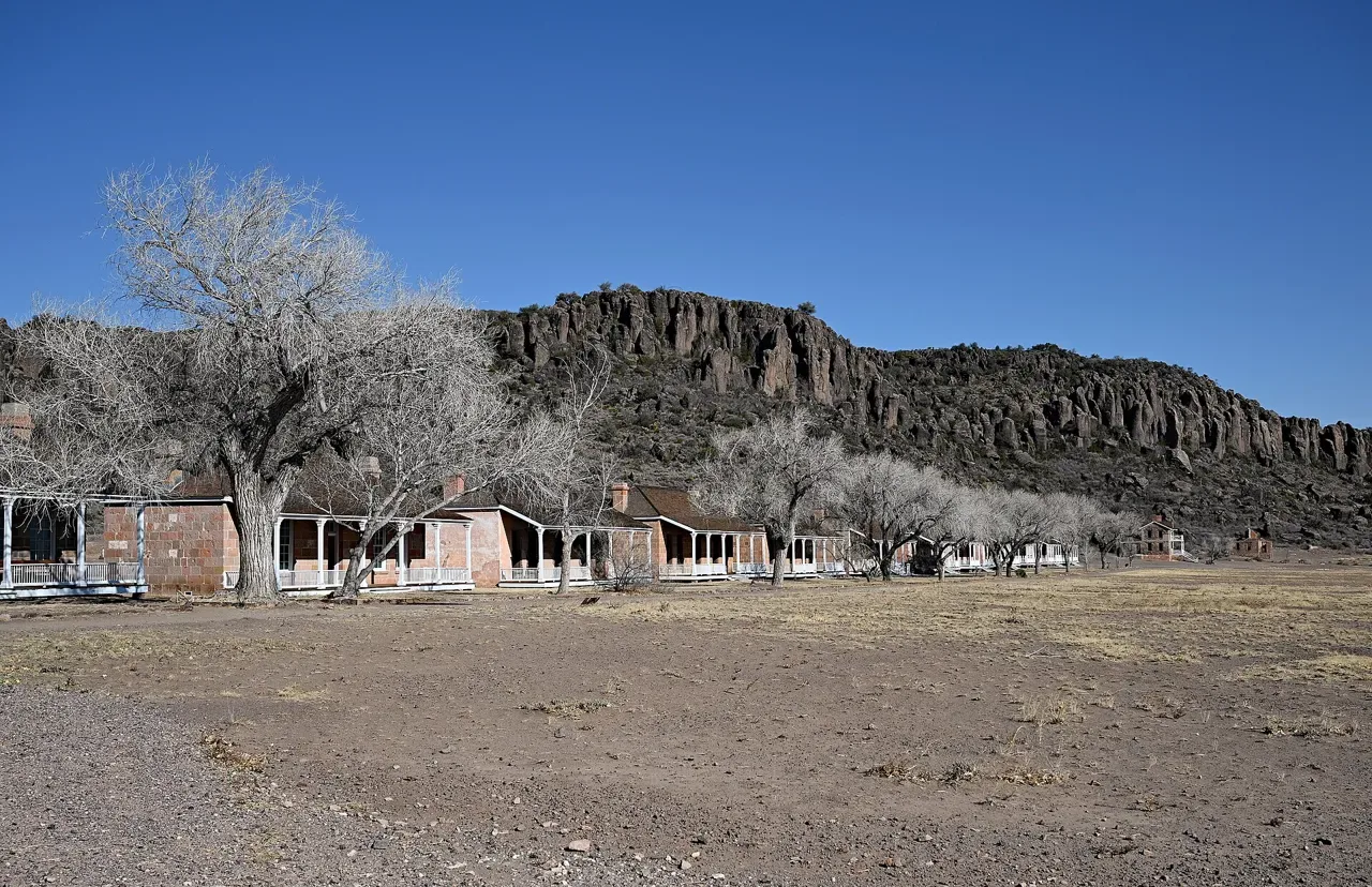 A long row of restored stone and adobe officers' quarters with white-columned porches and bare trees, at the foot of a cliff of tall rock columns under a deep blue sky, at Fort Davis, Texas