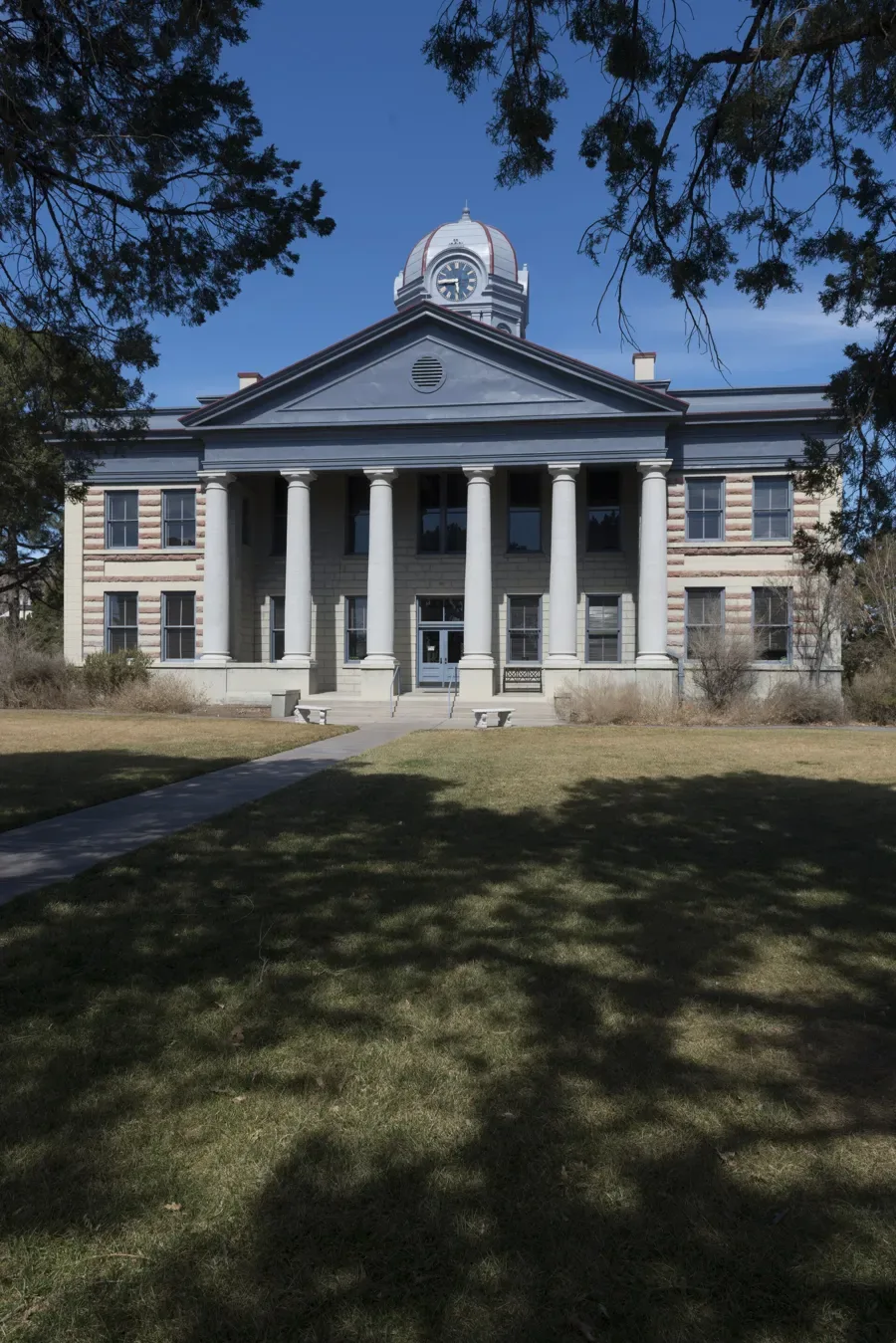 The Jeff Davis County Courthouse in Fort Davis, Texas, a two-story Classical Revival building with a pedimented portico of six tall columns, banded pink and tan stone walls, and a domed clock tower, seen across a shaded lawn