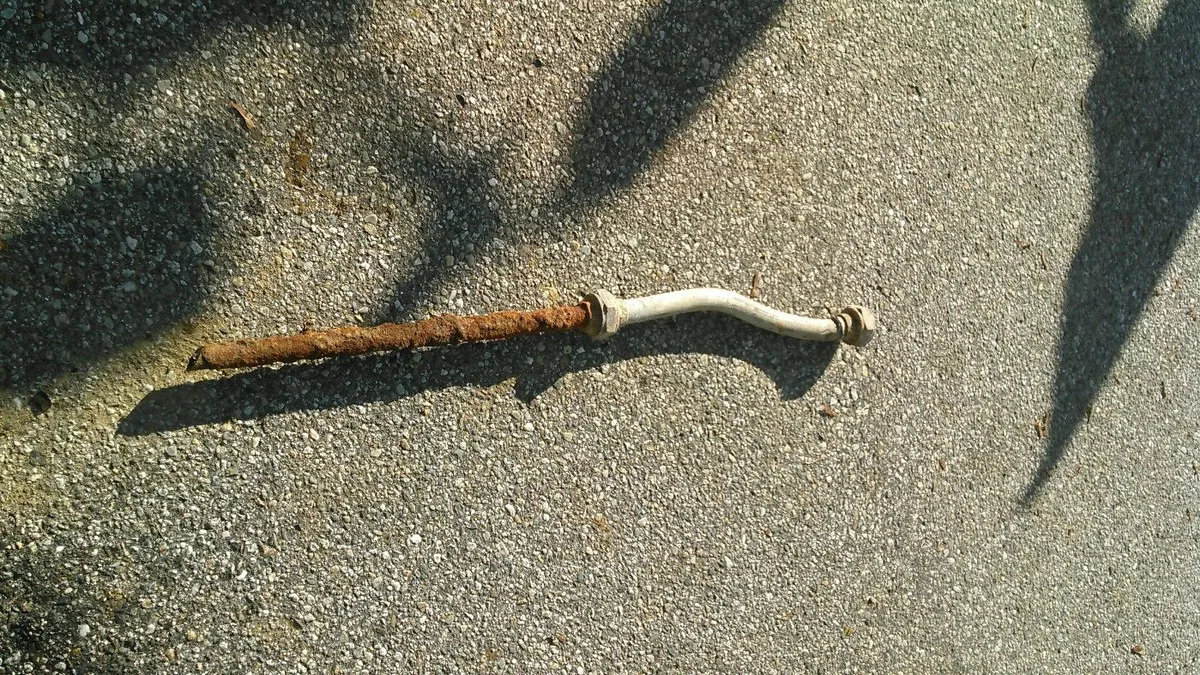 A rusted lead gooseneck pipe connected to a white flexible section, lying on pavement