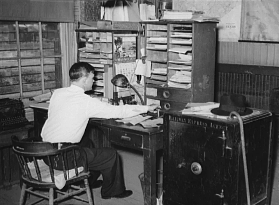 Black-and-white 1939 photograph of a man in a white shirt seated at a wooden desk in a cluttered office, with pigeonhole shelves, a calendar, a wall map and a large iron safe lettered Railway Express Agency beside the desk